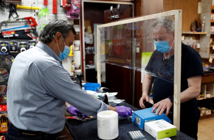 El trabajador de una ferretería atiende a un cliente con guantes, mascarilla y una pantalla protectora